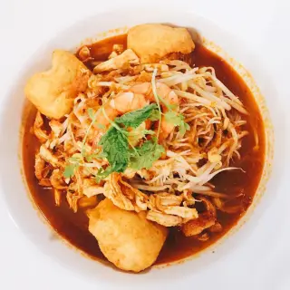 A bowl of laksa from above: tofu puffs, prawns, bean sprouts and coriander in a red curry broth