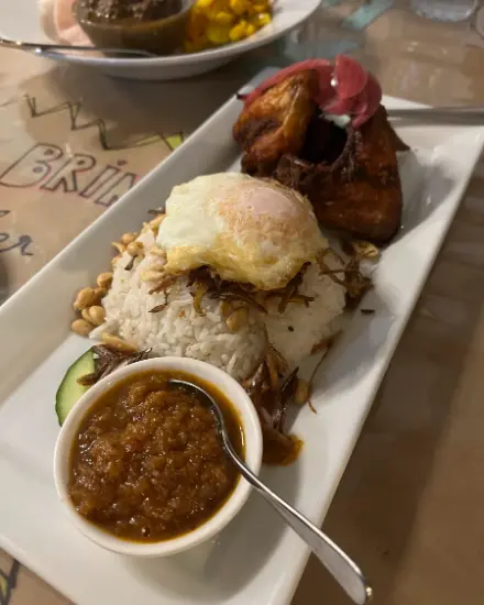Nasi lemak on a long white plate: coconut rice, fried egg, fried chicken, peanuts, anchovies, cucumber and a bowl of sambal